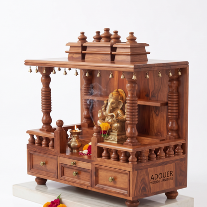 A wooden Ganesh idol display stand or miniature altar made of rich reddish-brown wood, featuring carved pillars, tiered shelves, small hanging bells, and three drawers. A brass Ganesh statue sits inside, with a diya and flowers arranged on and around it.